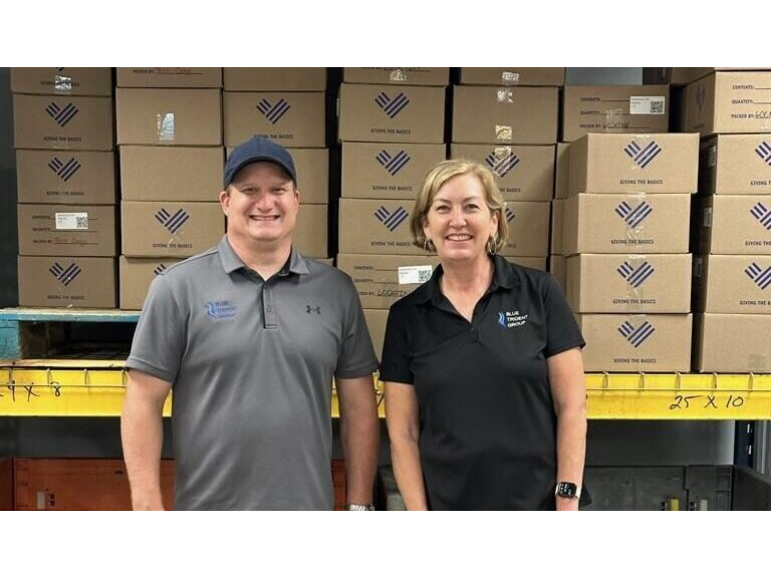 Kevin and Jill in front of a shelf of boxes in the Giving the Basics warehouse.